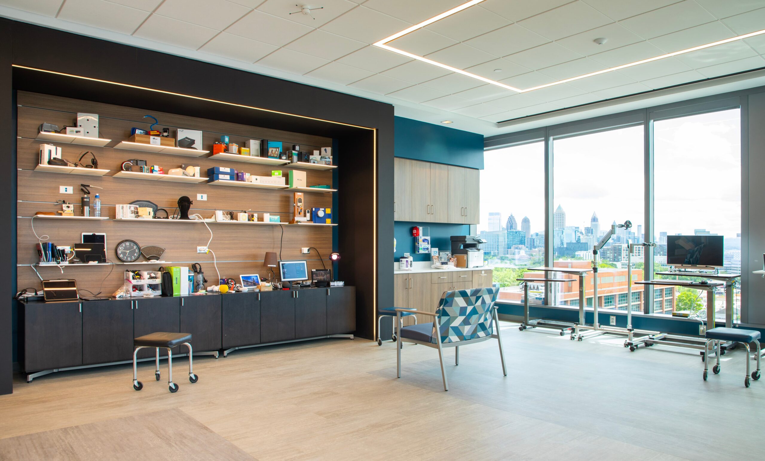 A modern, spacious office with large windows showing a city skyline, shelves filled with various gadgets, a patterned chair, cabinets, a small kitchen area, and a computer workstation.