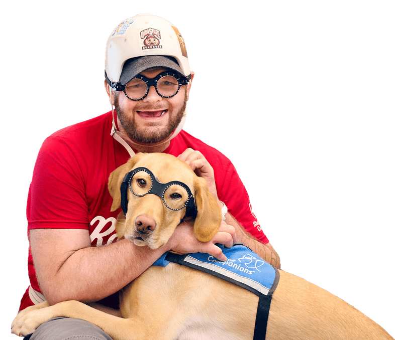 A smiling person wearing a cap, glasses, and a red shirt hugs a yellow dog. The dog, wearing a blue service vest, also has glasses on its face. Both are looking at the camera against a plain background.