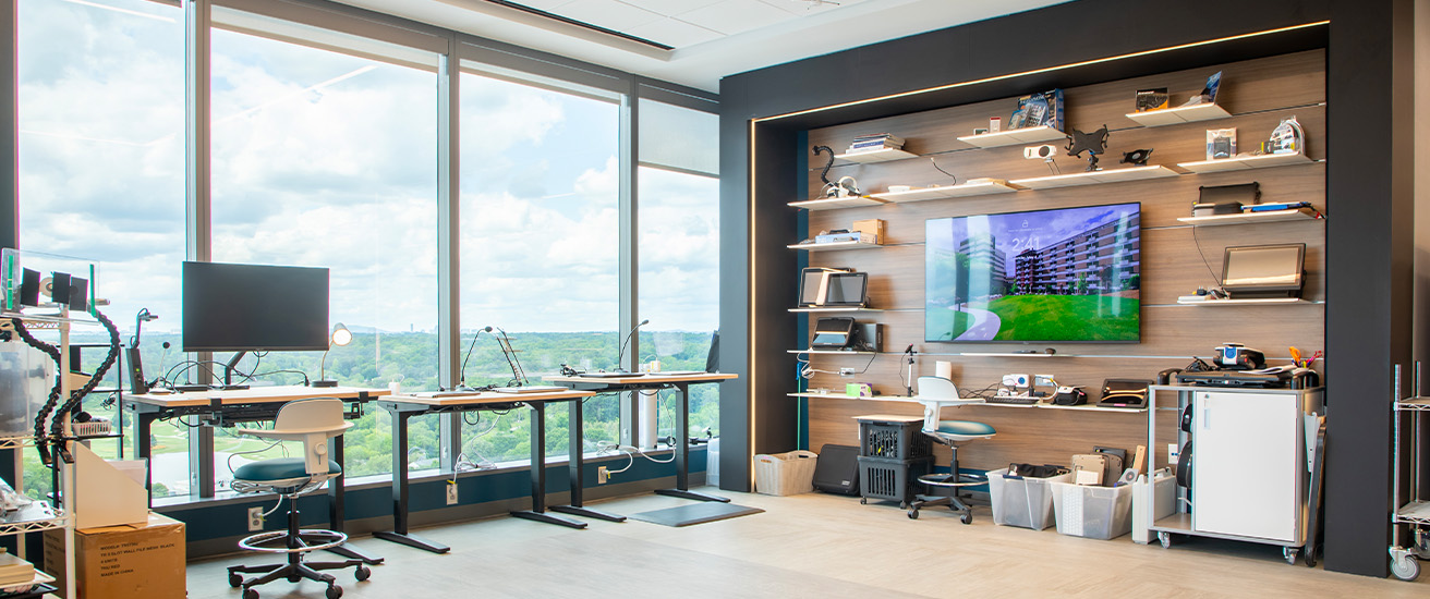 Modern office space with large windows, three desks and chairs, multiple computer monitors, and a wall display featuring shelves with various tech devices, a large screen showing a building, and bins under the shelves.
