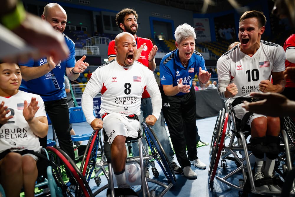 A group of ecstatic athletes in wheelchairs celebrates on a sports court. Clad in matching uniforms, they bask in joy as coaches and supporters join with wide smiles and claps.