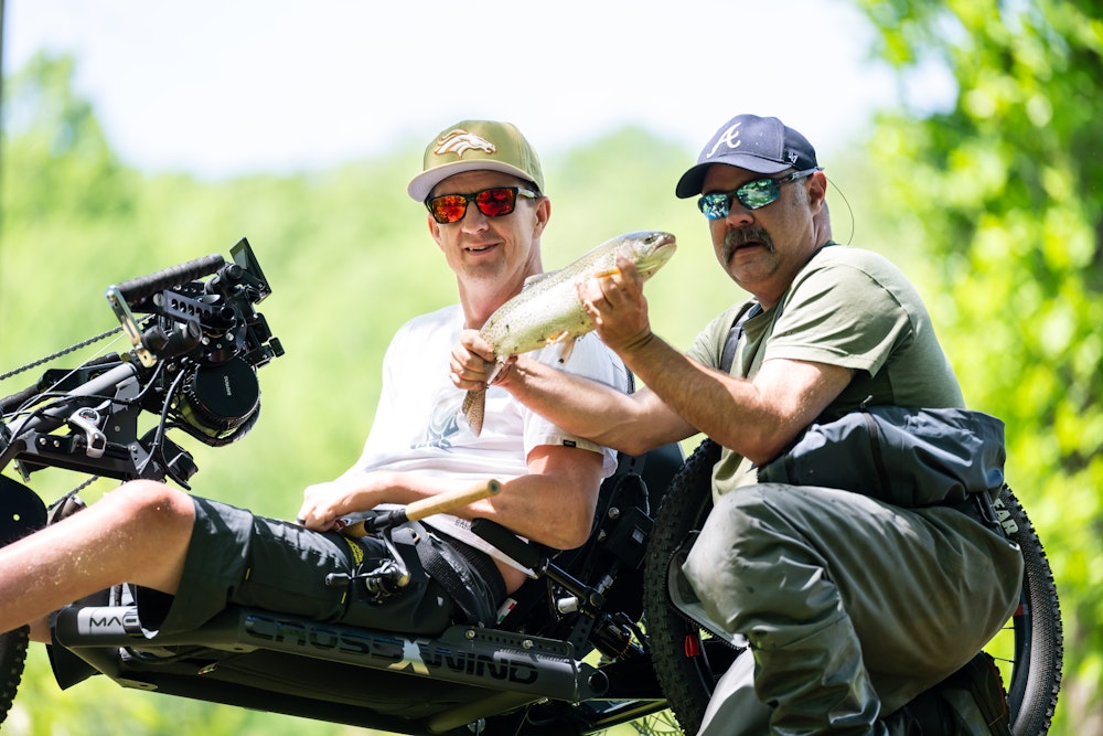 Two men outdoors, one holding a freshly caught fish and the other smiling beside him. The man on the left is sitting on an adaptive bicycle.