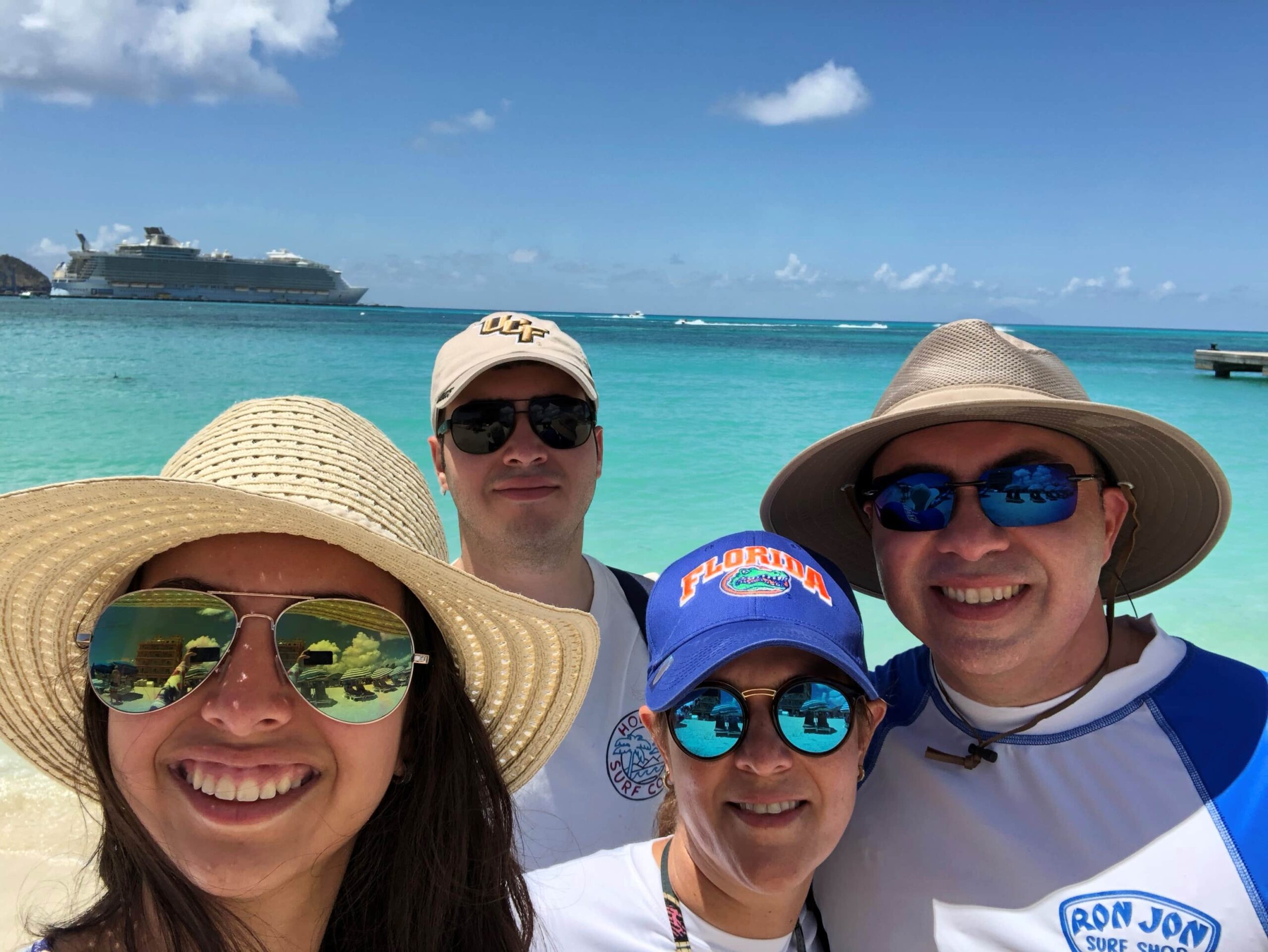 Four people in sunglasses pose happily on a sunny beach with turquoise water. A large cruise ship is visible in the background. They wear hats and beach attire, enjoying a clear day by the ocean.