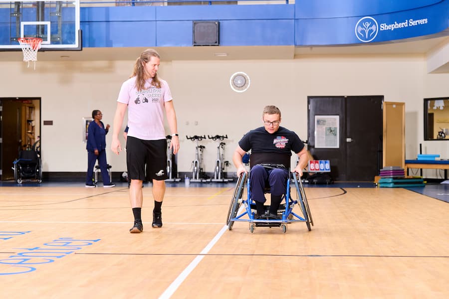 A man in a wheelchair moves across a gymnasium floor while another man walks beside him. A basketball hoop and exercise equipment are visible in the background.