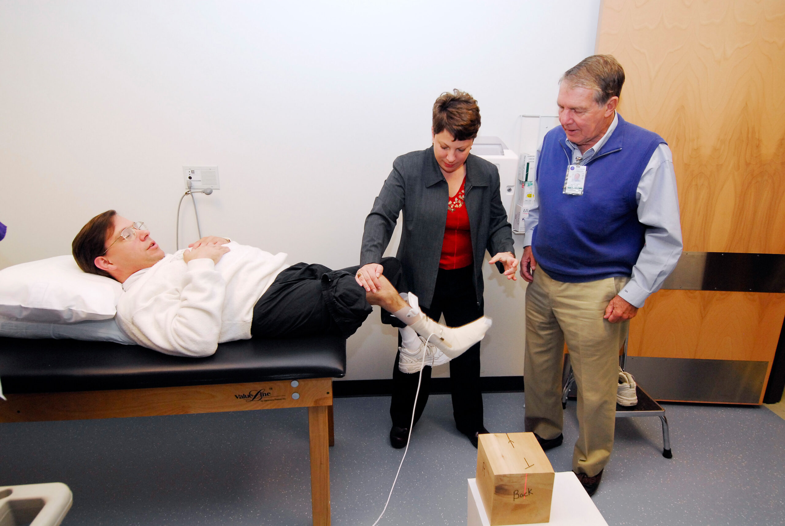 A man lies on a medical exam table while a woman checks his leg and Dr. David Apple observes, in a clinical setting.