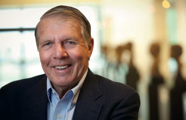 Dr. Apple smiling, wearing a dark suit jacket and light blue shirt stands indoors with a softly blurred background featuring abstract shapes and warm lighting.