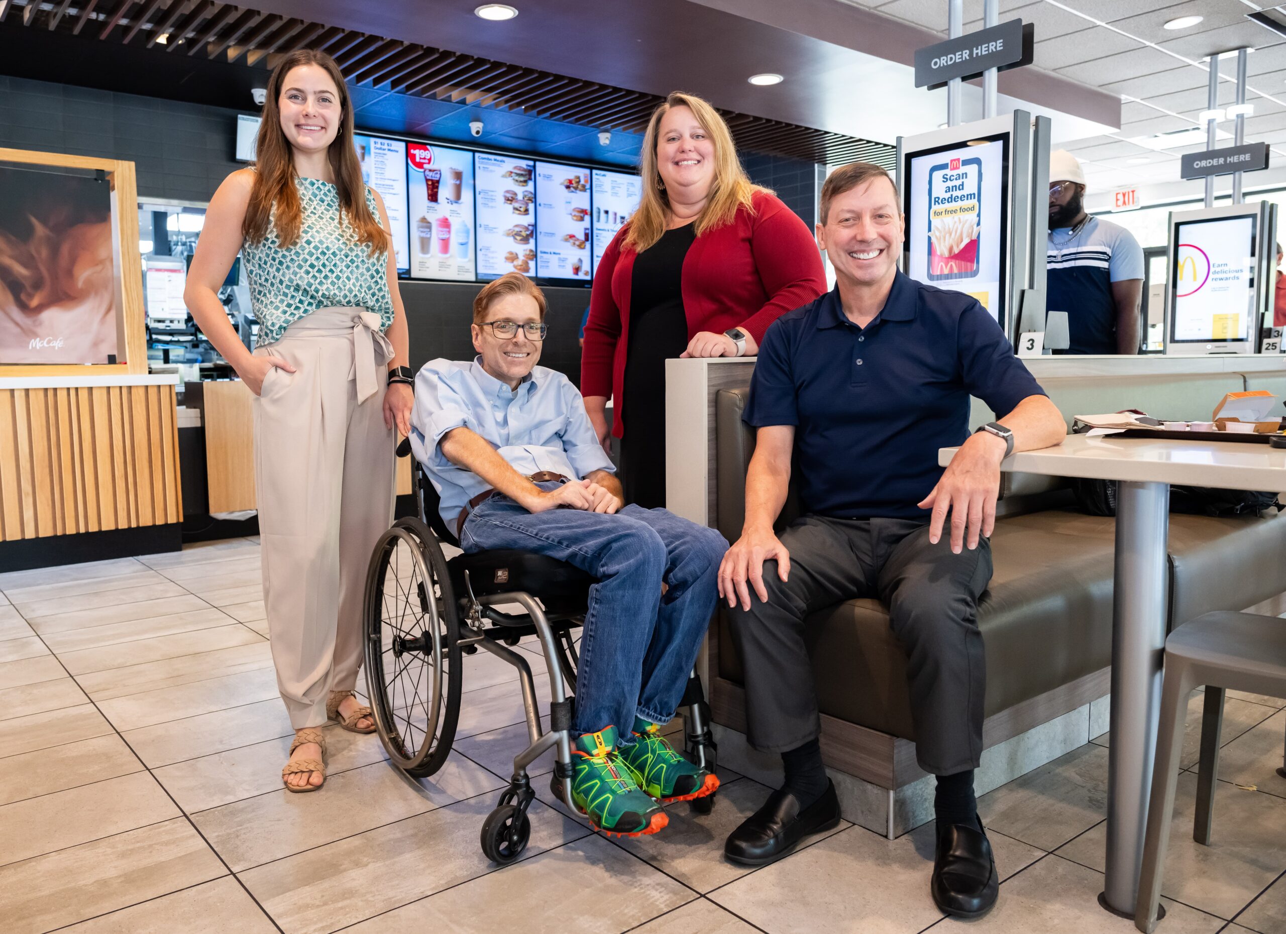 Four members from the AURC team pose and smile inside a fast-food restaurant. One man sits in a wheelchair, surrounded by two women standing and another man sitting on a booth seat. Digital menu boards are visible in the background.