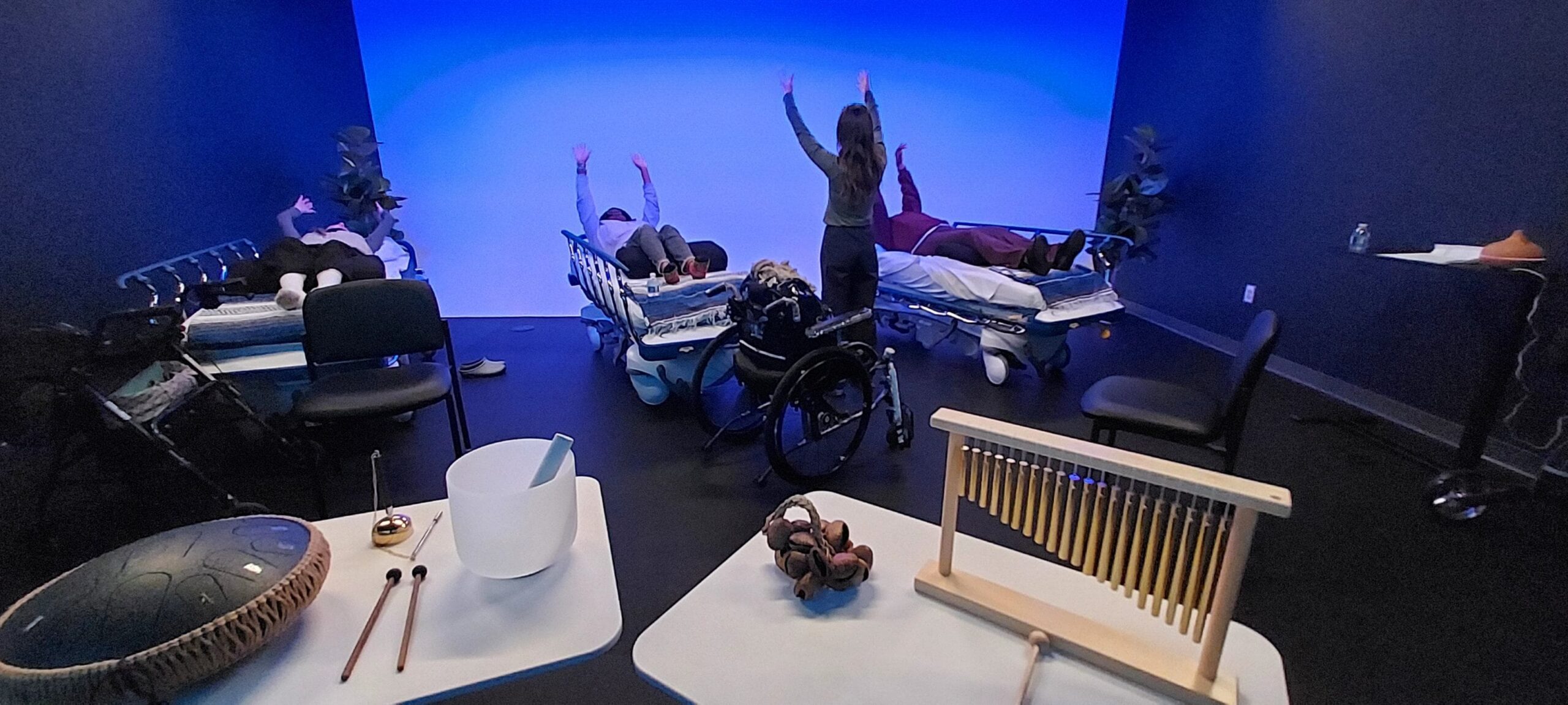 A group of people lies on beds in a dimly lit room with blue lighting while an instructor stands with arms raised. Various musical instruments are on tables in the foreground.