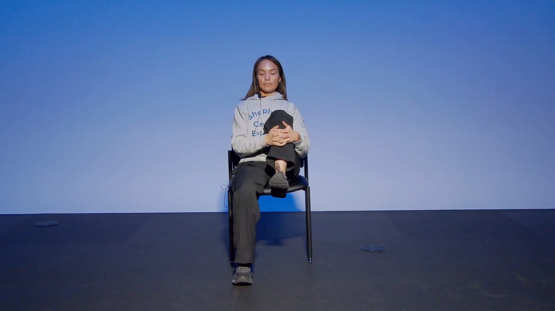 A person sits on a chair against a blue background, eyes closed, arms wrapped around their bent right knee, and left foot flat on the floor, appearing relaxed and thoughtful.