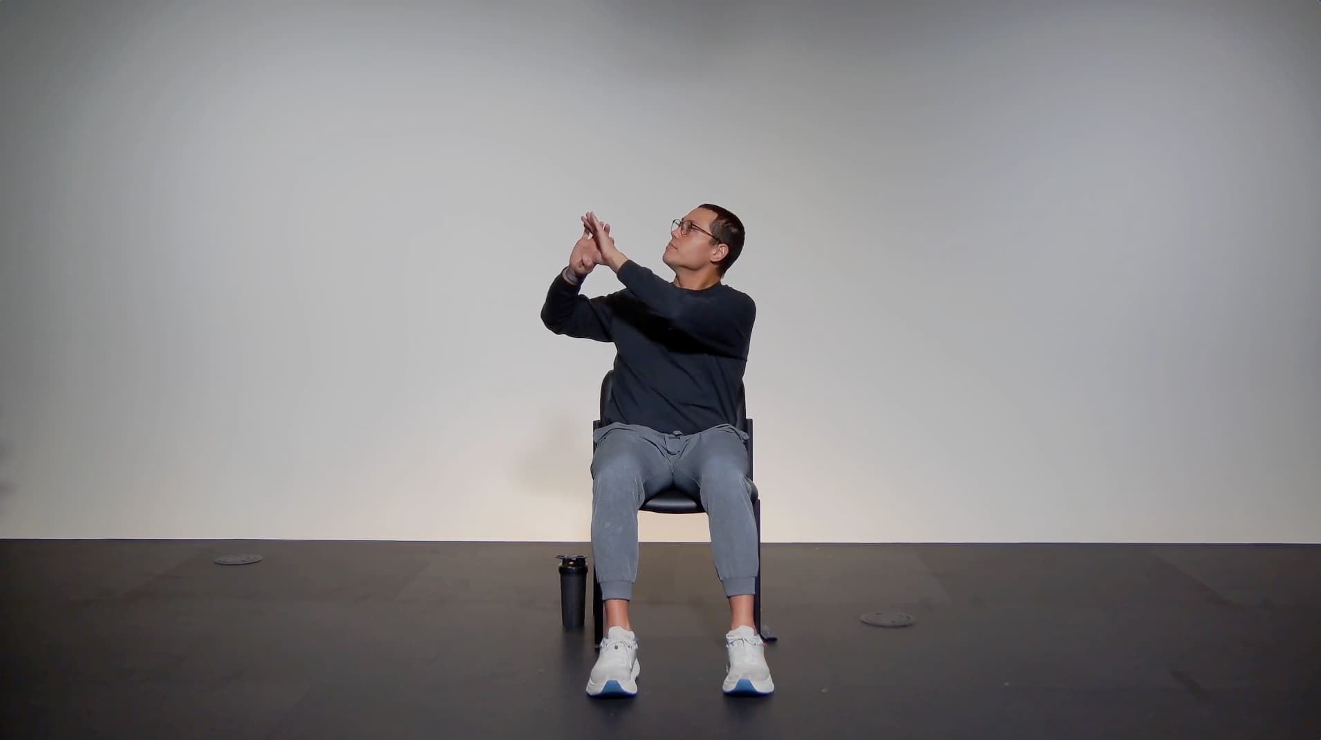 A person sits on a chair against a plain background, looking up and gesturing with their hands as if pointing or reaching towards something above. A water bottle is on the floor beside the chair.