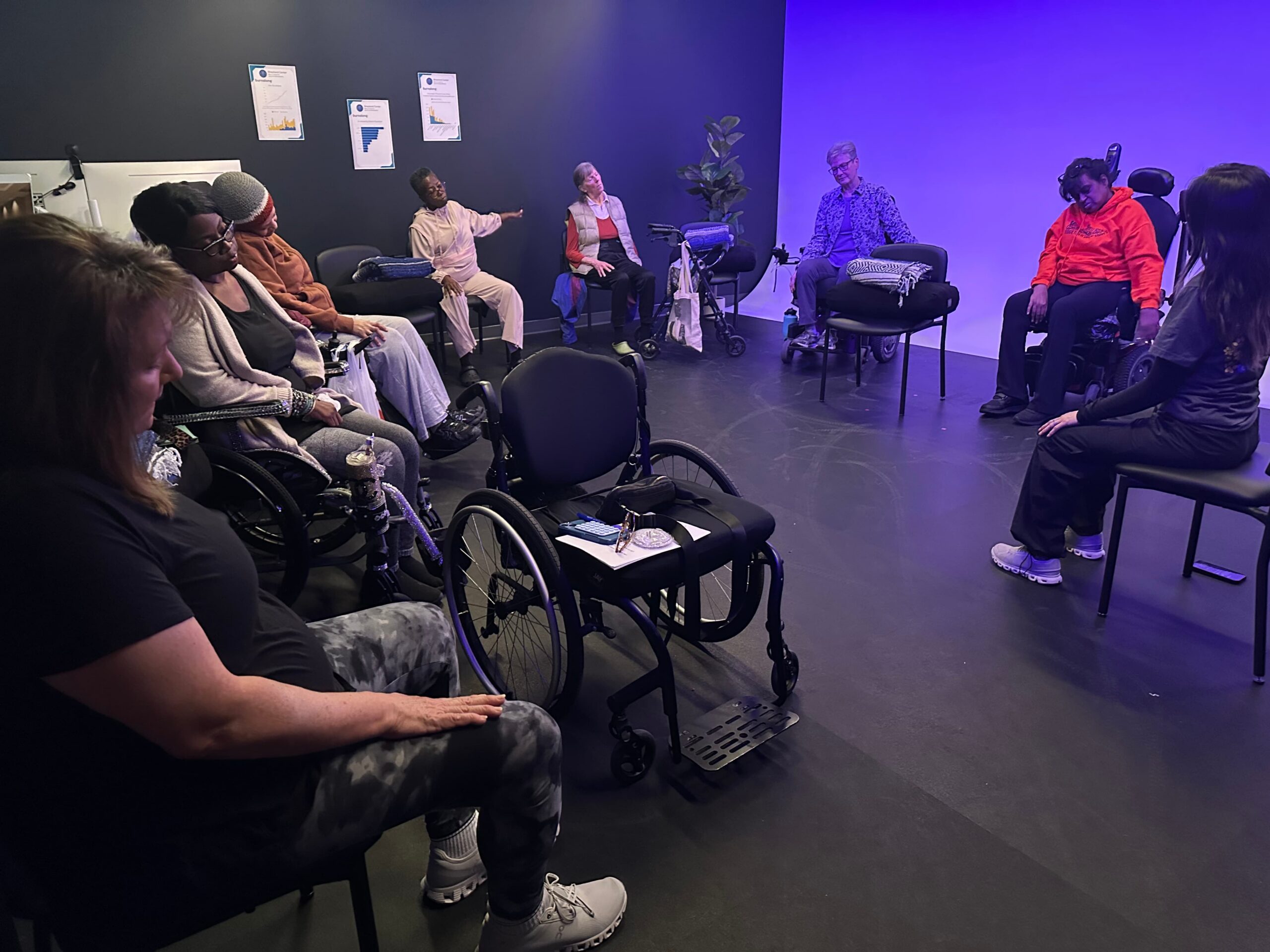 A group of people, some seated in wheelchairs and others in chairs, sit in a circle in a room with purple lighting, engaging in what appears to be a group discussion or therapy session.