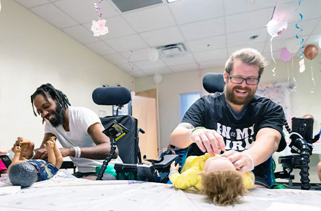 Two men in powered wheelchairs are smiling as they practice diapering baby dolls on a table. The room is decorated with colorful hanging ornaments, and they appear engaged and joyful in their activity.