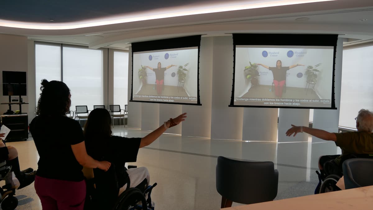 Two people in wheelchairs and a standing instructor follow an exercise video projected on two large screens in a bright, modern room. All participants are raising their arms, mirroring the movements in the video.