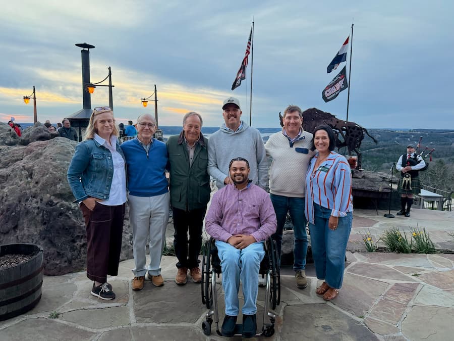 Seven adults, including one man in a wheelchair, pose together outdoors on a stone patio at sunset with flags, a sculpture, and a scenic landscape in the background.