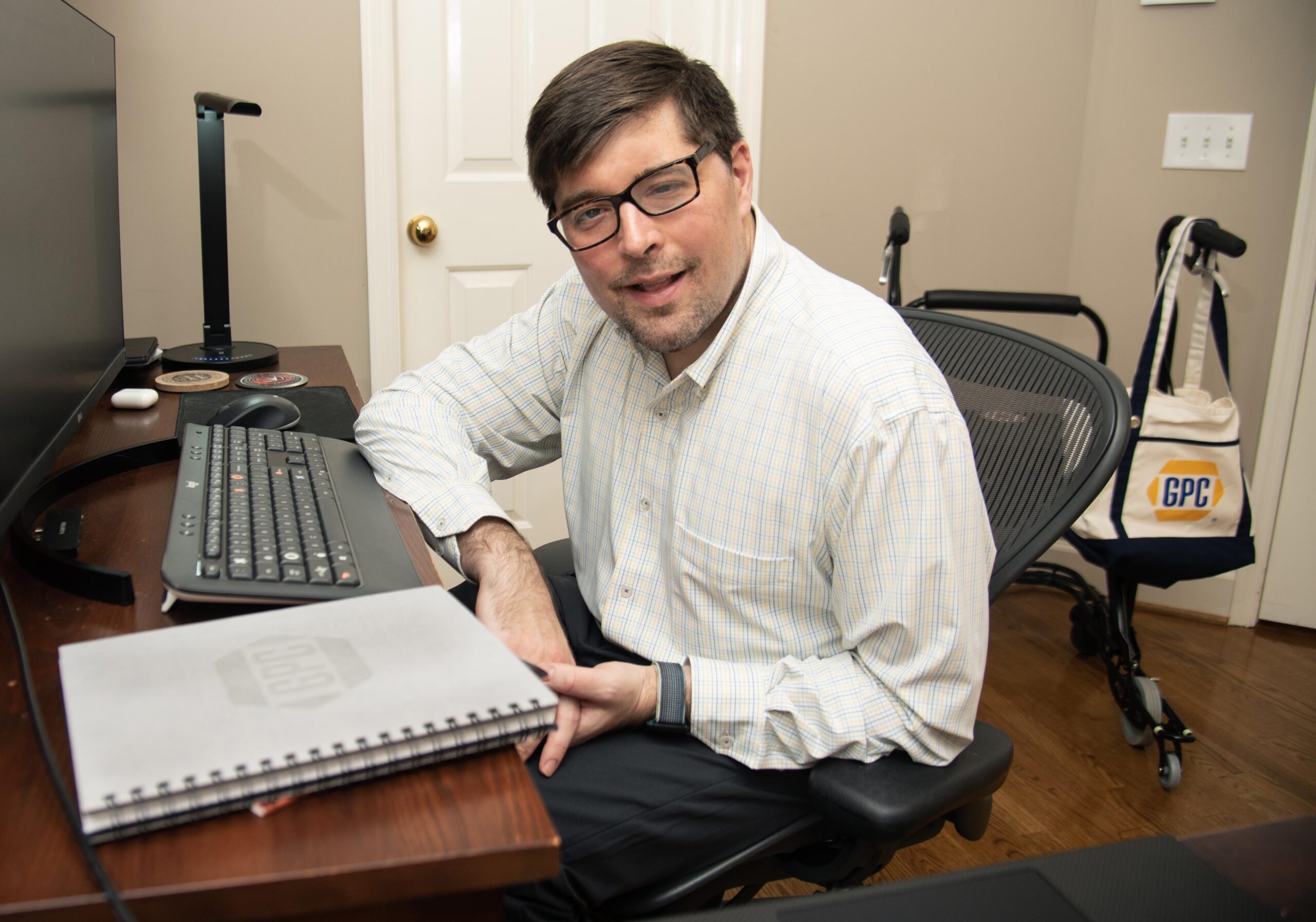 A person with short dark hair and glasses smiling, seated at a desk with a computer monitor and keyboard. A walker is visible in the background.