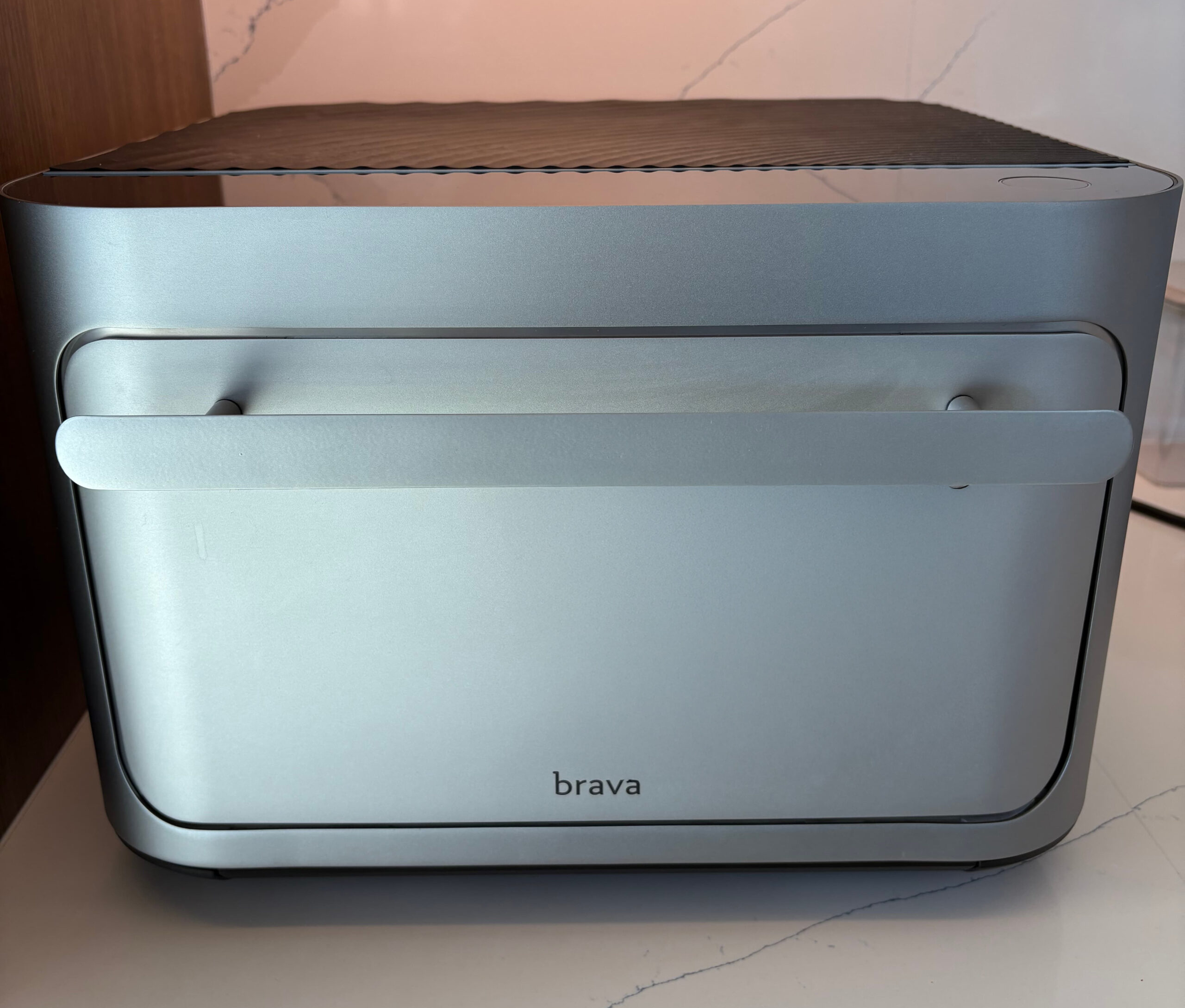A silver Brava countertop smart oven with a sleek, modern design sits on a white kitchen counter against a marble backsplash.