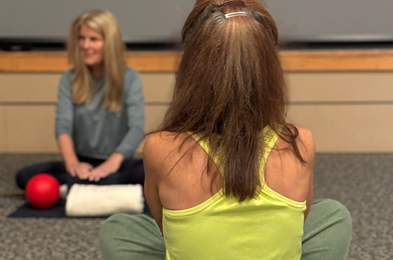 Two women are sitting on the floor in a room. One faces away, wearing a yellow tank top, while the other, in focus, faces the camera and wears a blue shirt. A red exercise ball and a white towel are on the floor between them.