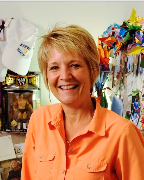 Cheryl Linden wears an orange shirt, standing in front of a colorful wall with paper cranes, notes, and memorabilia, including a wrestling action figure and a 