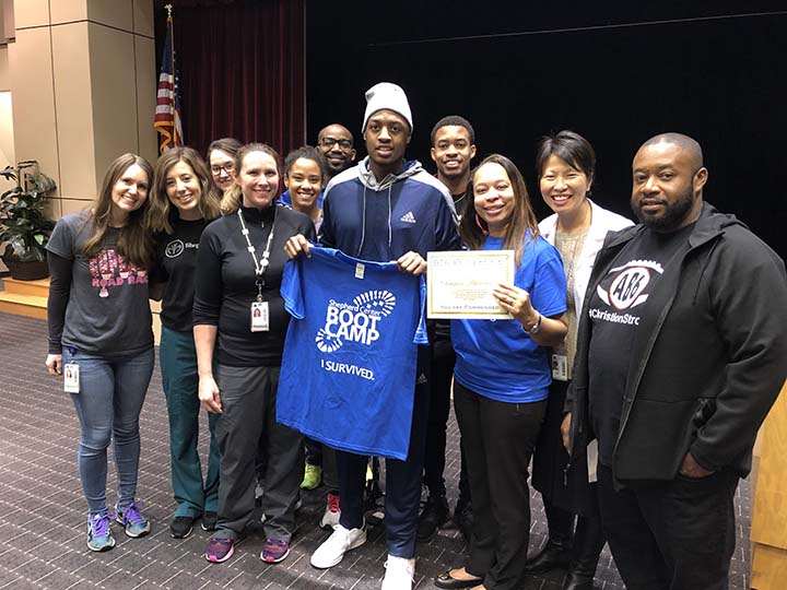 A group of people pose indoors, with one person in the center holding a blue “Boot Camp” t-shirt and another holding a certificate. They are smiling and standing on a dark carpeted floor near an American flag.