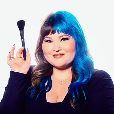 A woman with bright blue hair and bangs smiles while holding a makeup brush. She wears a dark top and has long, styled hair with brown and blue colors. The background is plain and bright.