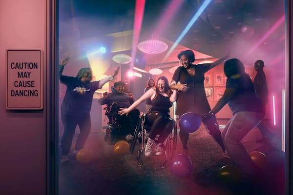 A diverse group of people, including wheelchair users, dance joyfully with balloons under colorful lights. A sign on the door reads, 