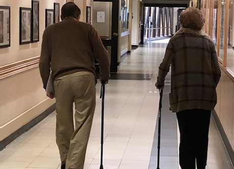 Dr. David Apple and Alana Shepherd walk down a hallway, each using a walking cane for support. The image is taken from behind as they move toward a well-lit corridor with signs overhead.