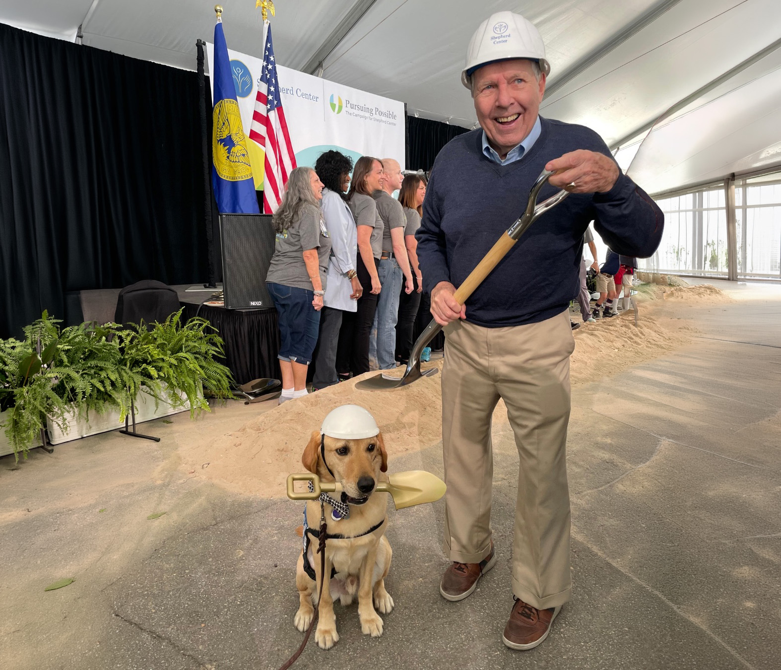 Dr. Apple wears a hard hat holds a shovel next to a yellow labrador facility dog also wearing a hard hat, inside a tent at the Arthur M. Blank Family Housing groundbreaking ceremony.