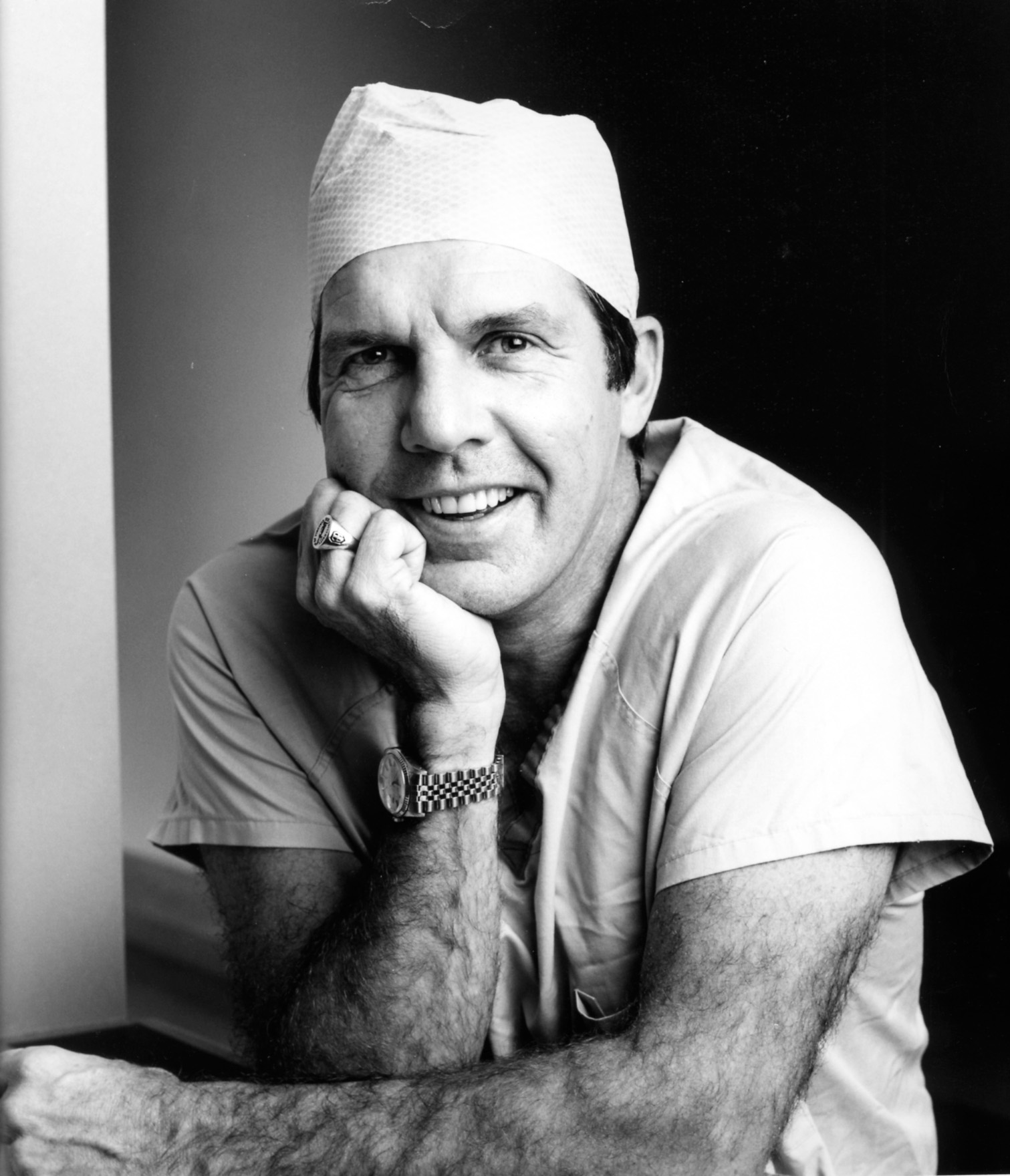Dr. David Apple in medical scrubs and a surgical cap smiles, resting his chin on his hand. He wears a wristwatch and a ring, and poses in a relaxed manner against a dark background. The photo is in black and white.
