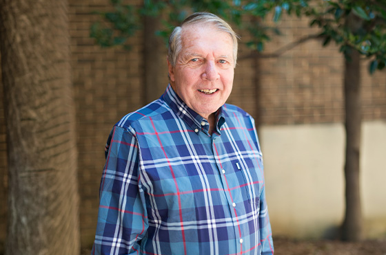 Dr. Apple wearing a blue plaid shirt stands outdoors, smiling at the camera. Behind him are trees and a brick wall.