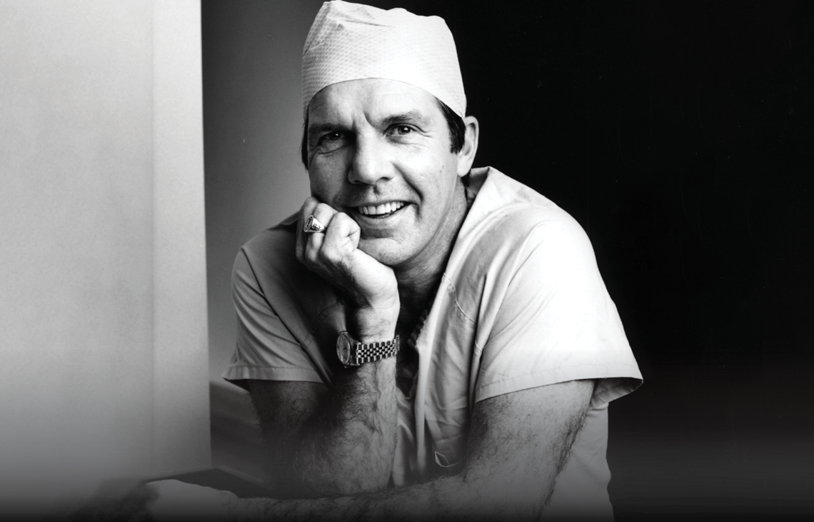 Dr. David Apple in medical scrubs and a surgical cap smiles, resting his chin on his hand. He wears a wristwatch and a ring, and poses in a relaxed manner against a dark background. The photo is in black and white.