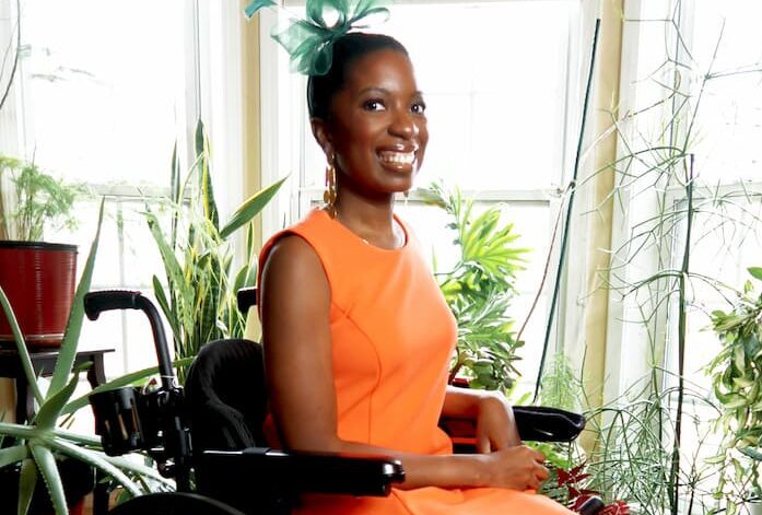 A woman in an orange dress and colorful sneakers sits in a wheelchair, smiling. She wears a green fascinator and is surrounded by houseplants in a sunlit room with a patterned red carpet.