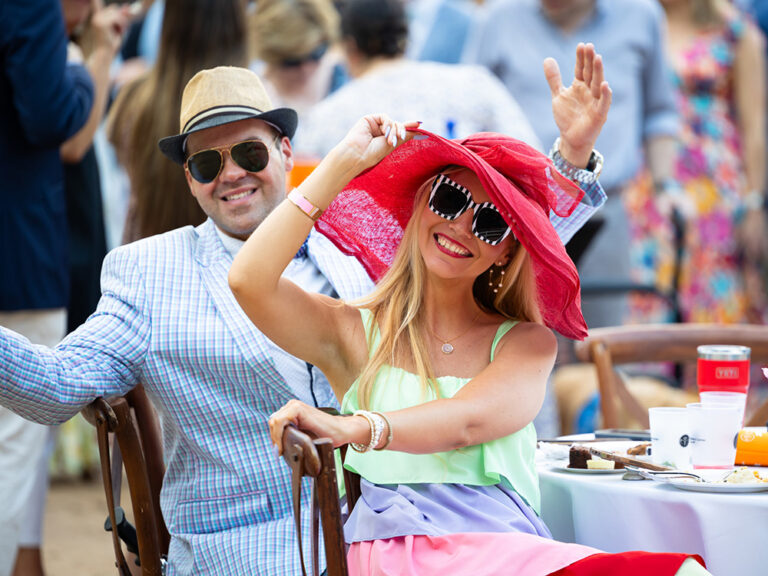 A man in a plaid blazer and a woman in a red sun hat and sunglasses smile at an outdoor event.