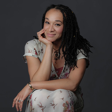 A woman with long, dark, braided hair and wearing a light floral dress sits with her chin resting on her hand, smiling softly against a dark gray background.