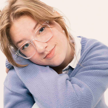 A young person with light brown hair and clear glasses rests their head on their arms, wearing a light purple sweater and smiling softly against a pale background.