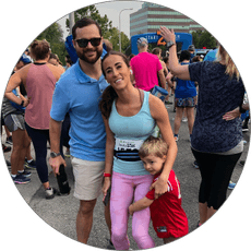 A man, woman, and young boy pose and smile at an outdoor race event with other participants gathered in the background. The boy hugs the woman’s leg, and everyone appears happy and relaxed.