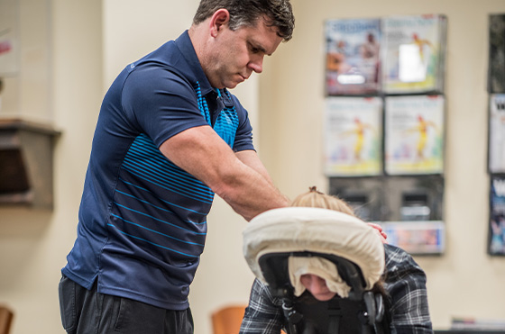 A massage therapist in a blue shirt and black pants is giving a seated massage to a person in a plaid shirt, who is resting on a massage chair. The setting appears to be an office or clinic with magazines on a shelf in the background.