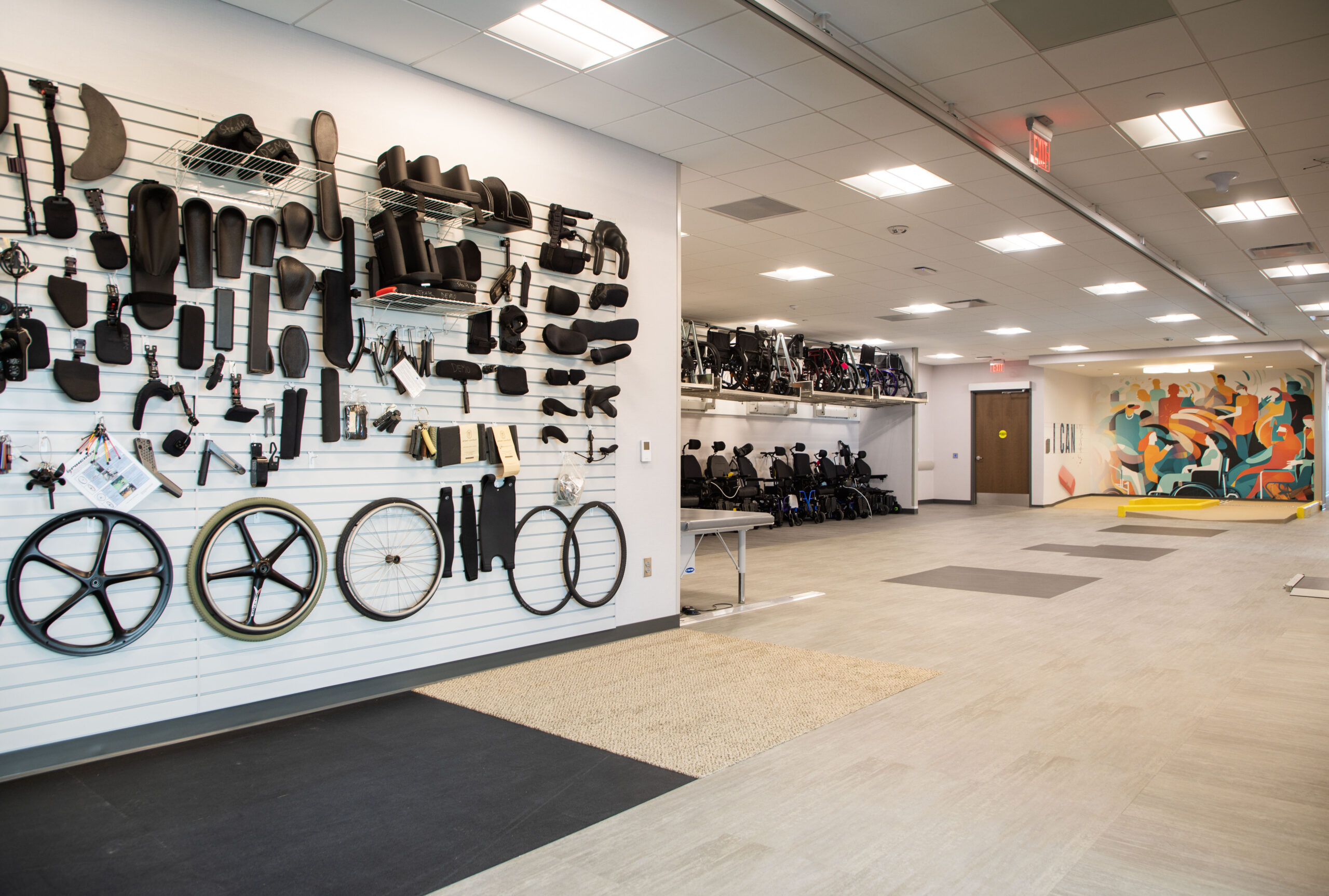 A spacious, modern room with wheelchair parts and accessories neatly displayed on wall racks, including wheels and seat cushions. Wheelchairs are lined up in the back, and a mural decorates the far wall.