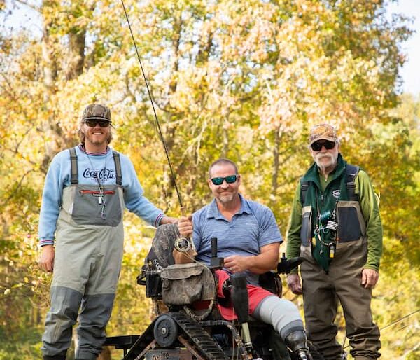 Three men outdoors by a wooded area: one in fishing gear, one seated in an all-terrain wheelchair with a fishing rod, and another in a green vest and waders. It’s a sunny day with trees in the background.