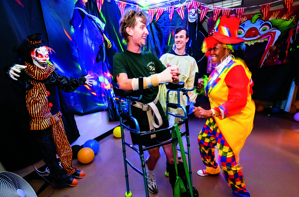 A person in colorful clown costume joyfully dance with a man using a walker at a lively, decorated indoor party with balloons, streamers, and a clown figure in the background.