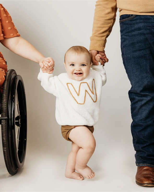 A smiling baby wearing a white sweater with a large “W” holds hands with two adults, one in a wheelchair and one standing, as they help the baby stand and walk.