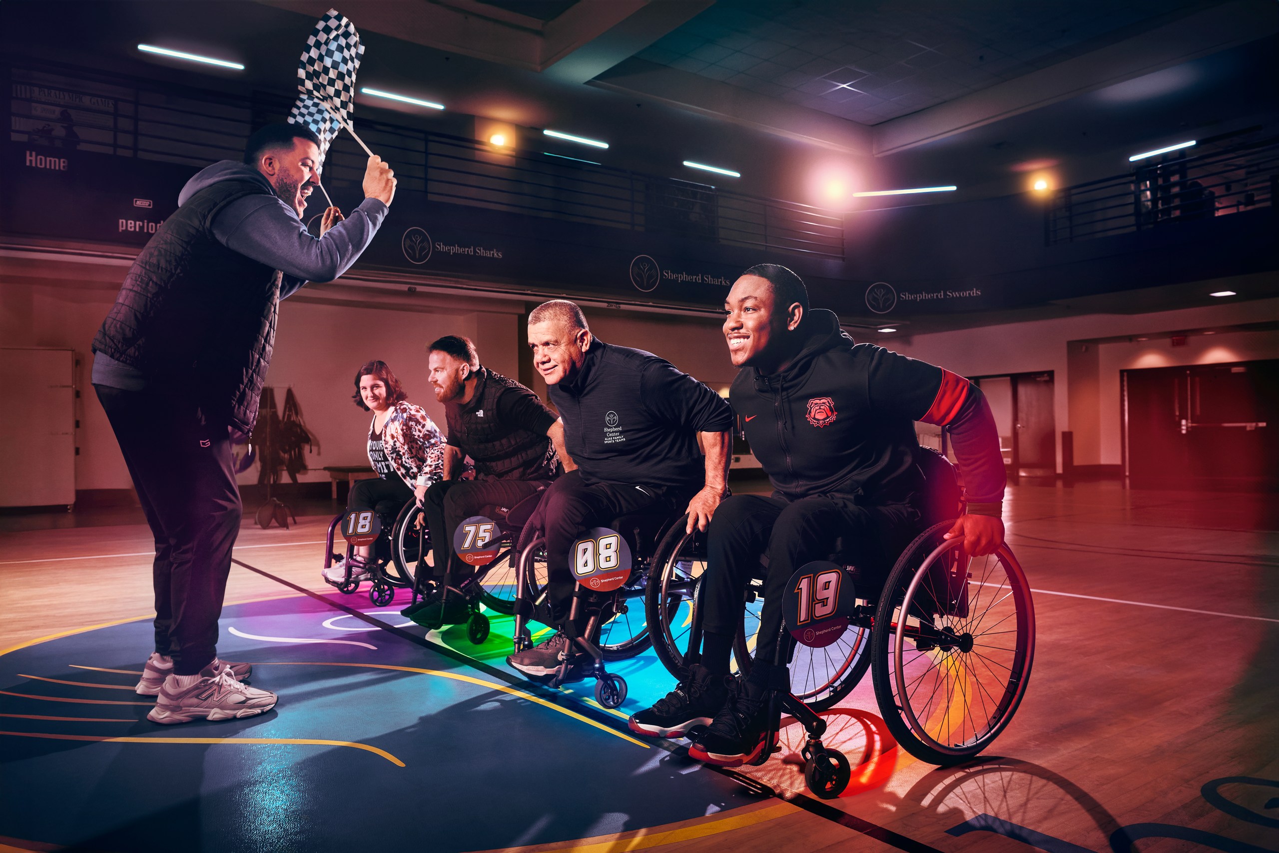 A coach waves a checkered flag as four wheelchair basketball players start a race on a gymnasium court.