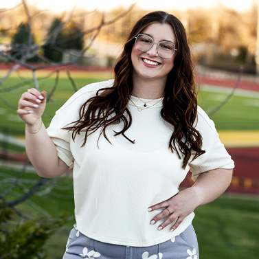 A woman with long brown hair and glasses smiles outdoors, wearing a white shirt and floral pants, standing in front of a sports field with blurred greenery and a running track in the background.