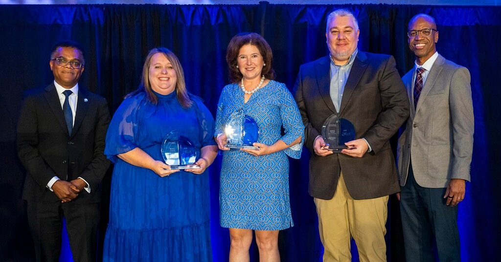 Five people stand on stage, dressed in formal attire, with a dark curtain and blue lighting in the background. Three people in the center hold glass awards and all are smiling at the camera.