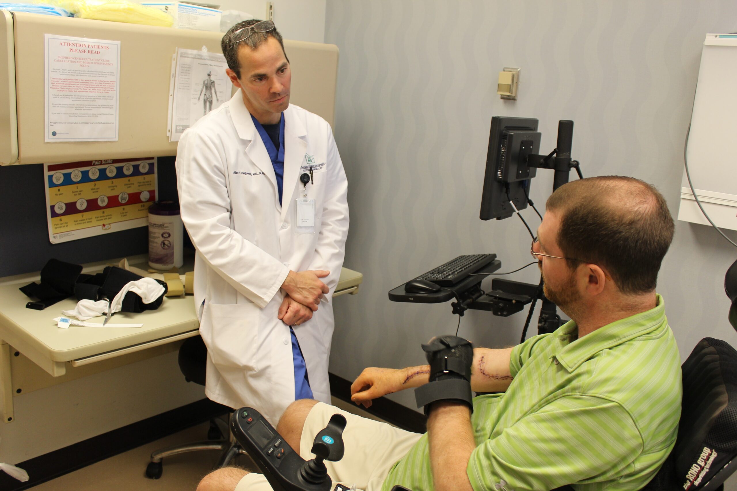 A doctor in a white coat attentively listens to a seated patient with a beard in a green shirt and adaptive gloves, who is in a wheelchair. They discuss upper extremity treatment and services at the Shepherd Center, surrounded by medical supplies and a computer in the office.