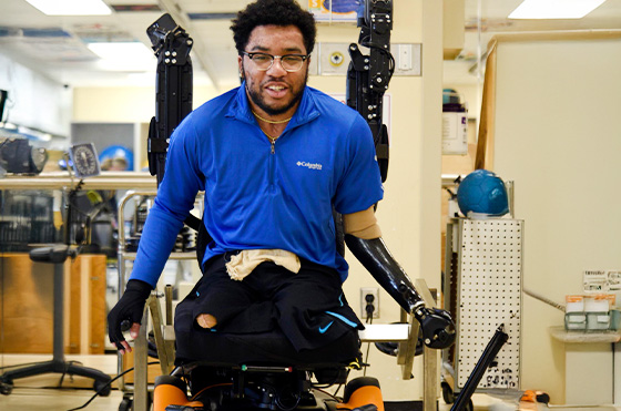 A man with prosthetic arms sits in a rehabilitation device, smiling. He wears glasses and a blue long-sleeve shirt. The background displays a therapeutic setting filled with equipment.