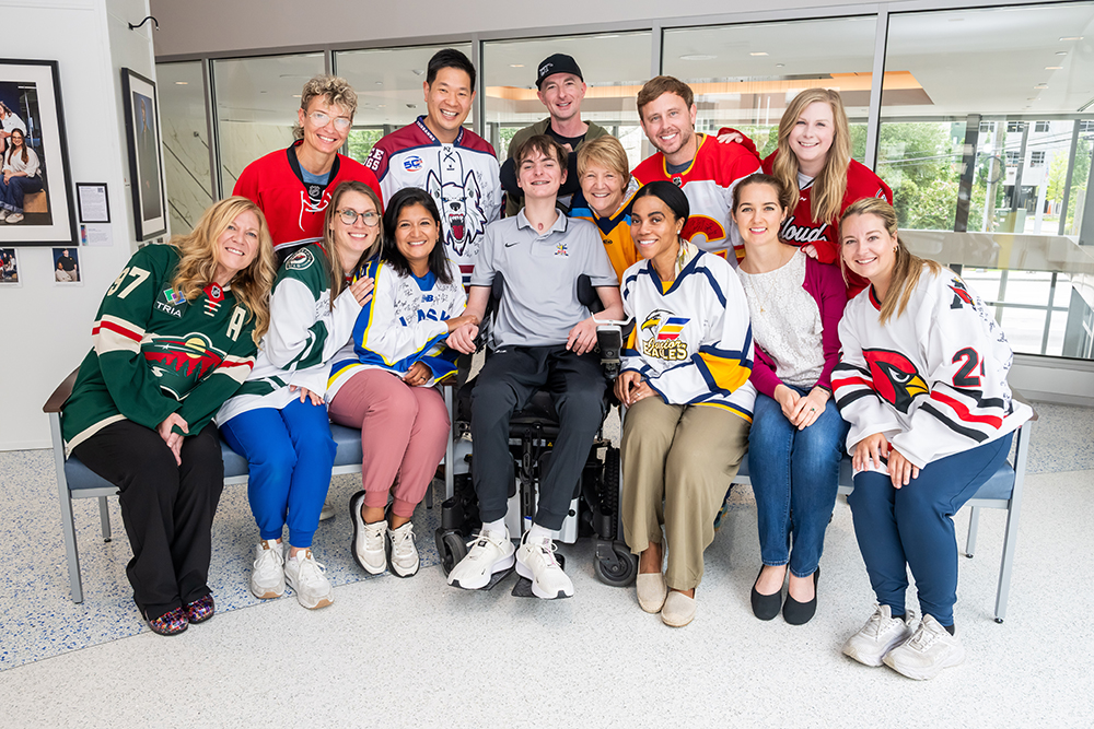 A group of twelve smiling people, some wearing hockey jerseys, pose together indoors. One person is seated in a wheelchair at the center, surrounded by friends standing and kneeling close, showing a supportive and cheerful atmosphere.