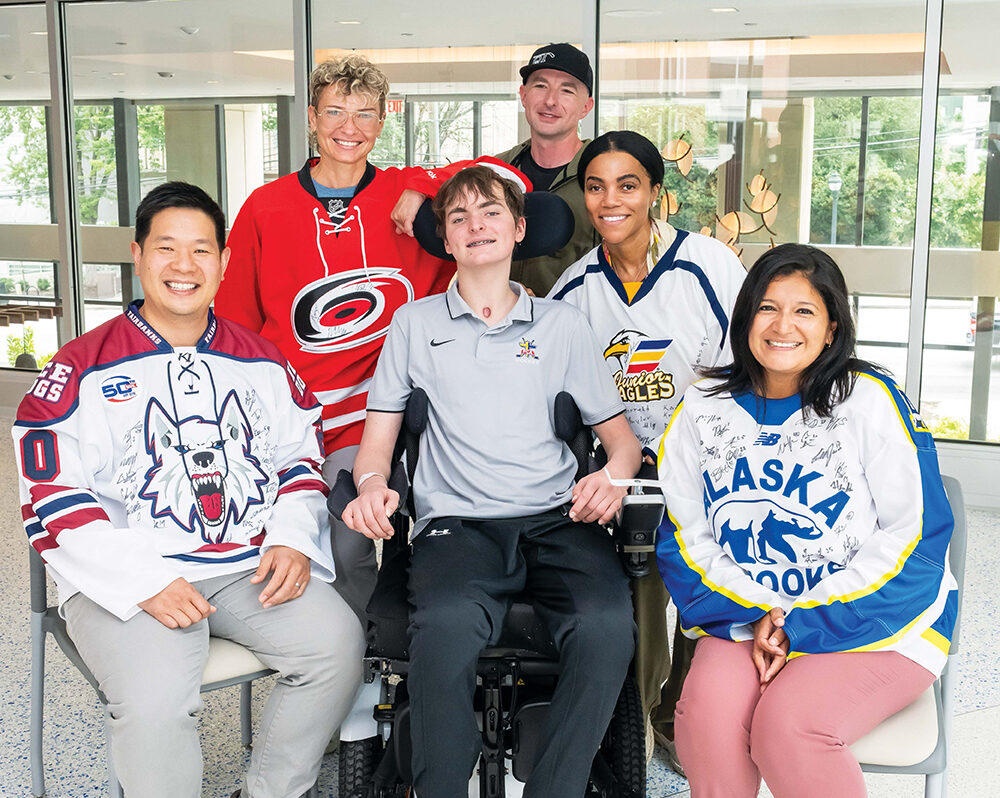 Six smiling people pose indoors, including a young man in a wheelchair at the center. The group wears various hockey jerseys and casual clothes, and they appear happy and supportive in a bright, modern setting.