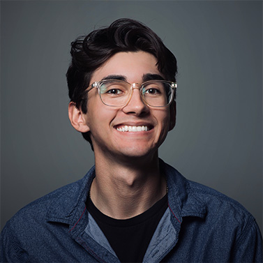 A young man with dark hair and glasses smiles broadly at the camera. He is wearing a dark denim shirt over a black t-shirt and stands against a plain, dark gray background.