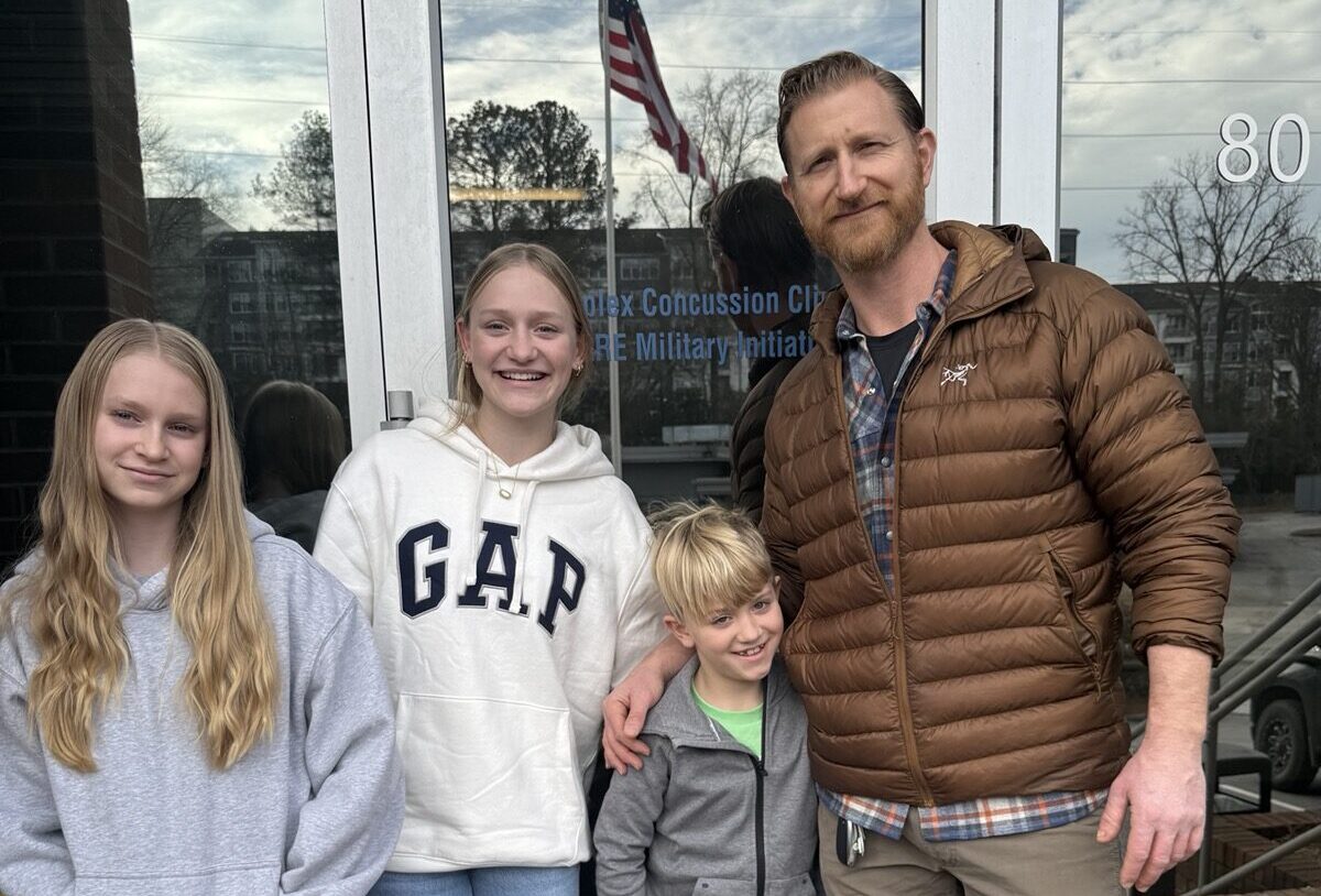 A man and three children stand smiling outside a glass door. The man wears a brown jacket. The children wear casual clothes. An American flag and trees are visible in the background.
