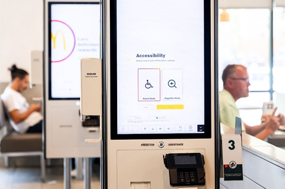 A McDonald's self-service kiosk displays accessibility options on its screen. In the background, two people are seated separately, one using a smartphone and the other sitting at a table.