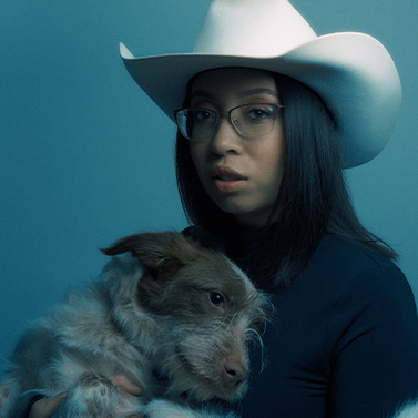 A woman wearing a white cowboy hat and glasses holds a small, scruffy dog. The background and lighting have a cool blue tone, giving the portrait a calm, moody atmosphere.
